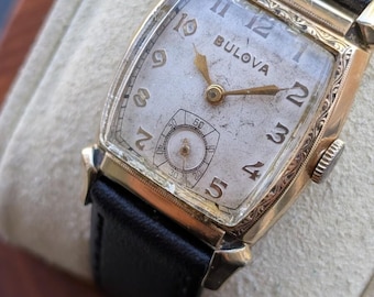 May include: A vintage Bulova wristwatch with a rectangular gold-tone case featuring intricate engravings on the bezel. The off-white dial displays black Arabic numerals, gold-tone hands, and a small sub-dial for seconds. It has a black leather strap.