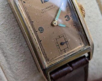 May include: A rectangular wristwatch with a gold-tone case and a brown leather strap. The dial is a muted tan colour featuring the text Roamer Standard, black Arabic numerals at 12, 3, and 9, a small seconds sub-dial, and light blue hands.