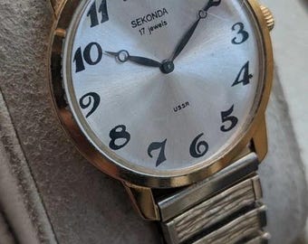 Könnte beinhalten: Eine goldfarbene Sekonda Armbanduhr mit silbernem Zifferblatt im Sonnenschliff, schwarzen arabischen Ziffern und schwarzen Zeigern. Das Zifferblatt trägt die Aufschrift 'SEKONDA', '17 jewels' und 'USSR'. Sie hat ein strukturiertes, flexibles Metallarmband.