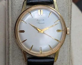 Könnte beinhalten: Eine Avia Staccato Armbanduhr mit rundem goldfarbenem Gehäuse, weißem Zifferblatt mit goldfarbenen Stundenmarkierungen und Zeigern sowie einem schwarzen Lederarmband. Das Zifferblatt zeigt den Text AVIA Staccato und Swiss Made am unteren Rand.