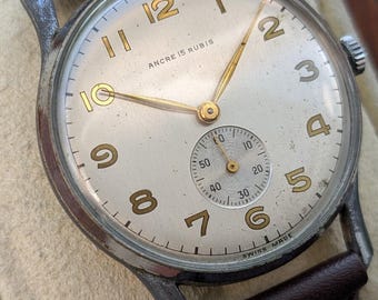 May include: A vintage wristwatch with a silver-toned metal case and a dark brown leather strap. The off-white dial features gold-coloured Arabic numerals, gold hands, and a sub-dial at the six o'clock position. Text on the dial reads ANCRE 15 RUBIS and SWISS MADE.