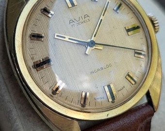 Könnte beinhalten: Eine Avia 17 Jewels Armbanduhr mit einem goldfarbenen runden Gehäuse und einem strukturierten champagnerfarbenen Zifferblatt. Die Uhr verfügt über goldfarbene Stundenmarkierungen mit schwarzen Akzenten, dünne goldene Zeiger und ein braunes Lederarmband. Das Zifferblatt ist mit Incabloc und Swiss Made beschriftet.