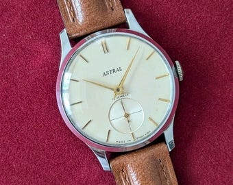 May include: A vintage Astral wristwatch with a silver-tone round case, a cream-coloured dial, gold-tone hands, and a sub-dial for seconds. The watch features the text Astral, 17 Jewels, and Made in England on the face, paired with a brown leather strap.
