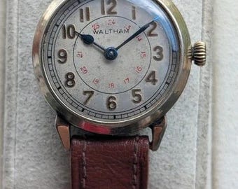 Könnte beinhalten: Eine Vintage-Armbanduhr von Waltham mit rundem goldfarbenem Gehäuse, cremefarbenem Zifferblatt mit schwarzen arabischen Ziffern und einer roten 24-Stunden-Skala. Die Uhr hat dunkelblaue Zeiger und ein strukturiertes braunes Lederarmband.
