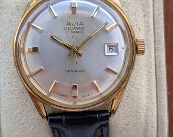 May include: A gold-tone wristwatch with a silver sunburst dial, featuring the text AVIA OLYMPIC 17 JEWELS and INCABLOC. The watch has gold-tone hour markers, a date window at the three o'clock position, and a black leather strap with a crocodile texture.