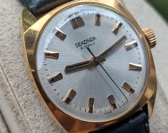 Könnte beinhalten: Eine goldfarbene Sekonda Armbanduhr mit silbernem Sonnenschliff-Zifferblatt, goldfarbenen Indizes und Zeigern. Die Uhr hat ein schwarzes Lederarmband und trägt die Aufschrift "SEKONDA 19 JEWELS" sowie "MADE IN USSR" auf dem Zifferblatt.