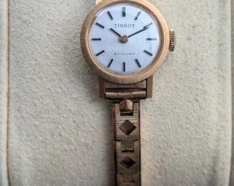 May include: A gold-tone Tissot Stylist wristwatch featuring a round white dial with black hour markers, thin black hands, and a textured metal link bracelet with geometric cutouts. The watch face displays the Tissot brand name and Stylist text.