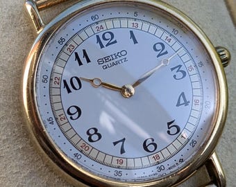 May include: A gold-tone Seiko Quartz wristwatch with a white dial, black Arabic numerals, and a gold mesh strap. The dial features a secondary 24-hour inner ring, gold-tone hands, and the text Seiko Quartz printed in black below the 12 o'clock position.