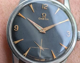 Könnte beinhalten: Eine Vintage-Armbanduhr von Omega mit einem runden silberfarbenen Gehäuse und einem schwarzen Zifferblatt. Das Zifferblatt zeigt goldfarbene Stundenmarkierungen, goldene Zeiger sowie die Ziffern 3, 9 und 12. Mit schwarzem Lederarmband.