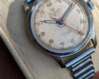 May include: A vintage Reconvilier wristwatch with a weathered, off-white dial, silver-tone Arabic numerals, and a red second hand. The watch features a metallic expansion band, a circular silver-tone case, and text reading Reconvilier, Ancre 17 Rubis, Antimagnetic, and Incabloc.