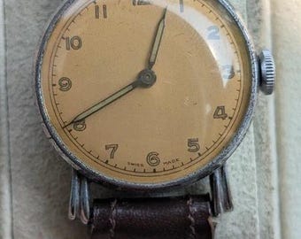 Könnte beinhalten: Eine Vintage-Armbanduhr mit rundem silberfarbenem Gehäuse und beigefarbenem Zifferblatt mit schwarzen arabischen Ziffern. Die Uhr hat dünne dunkle Zeiger und ein dunkelbraunes Lederarmband. Unten steht der Text "SWISS MADE" in kleiner Schrift.