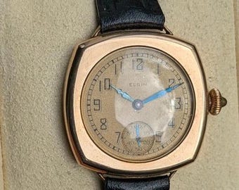 Könnte beinhalten: Eine Vintage-Armbanduhr von Elgin mit einem goldfarbenen, kissenförmigen Gehäuse, einem strukturierten beigen Zifferblatt und schwarzen arabischen Ziffern. Die Uhr hat leuchtend blaue Zeiger, ein kleines Sekunden-Hilfszifferblatt und ein schwarzes Lederarmband.