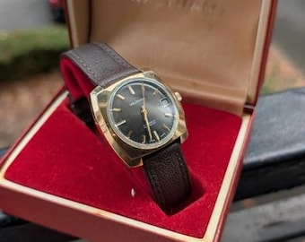 Könnte beinhalten: Eine goldfarbene Waltham-Armbanduhr mit schwarzem Zifferblatt, Datumsanzeige und dunkelbraunem, strukturiertem Lederarmband, präsentiert in einer rot ausgekleideten Schatulle mit dem eingeprägten Markennamen Waltham auf dem beigen Innendeckel.