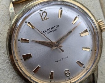 May include: A gold-tone wristwatch with a round silver dial, featuring the text Excalibur, 17 Jewels, and Incabloc. The watch has gold-tone hour markers, gold hands, and a flexible gold-tone expansion band. The dial includes the numbers 12 and 6.