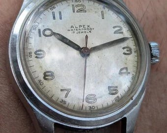 Könnte beinhalten: Eine wasserdichte Vintage-Armbanduhr von Alpex mit einem silberfarbenen Metallgehäuse und einem passenden Mesh-Armband. Das cremefarbene Zifferblatt zeigt schwarze arabische Ziffern, einen zentralen Sekundenzeiger und den Aufdruck Alpex Waterproof 17 Jewels.