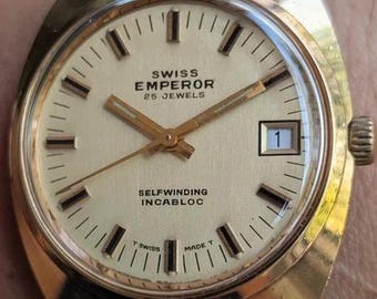 Könnte beinhalten: Eine goldfarbene Swiss Emperor Armbanduhr mit cremefarbenem Zifferblatt, goldenen Stundenmarkierungen und goldenen Zeigern. Die Uhr verfügt über ein Datumsfenster auf der Drei-Uhr-Position, ein braunes Lederarmband und den Schriftzug Swiss Emperor 25 Jewels.