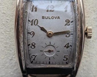 May include: A rose gold-tone Bulova wristwatch with a rectangular case and a textured metal link bracelet. The silver dial features black Arabic numerals, a small sub-dial for seconds at the bottom, and two gold-tone hands for hours and minutes.