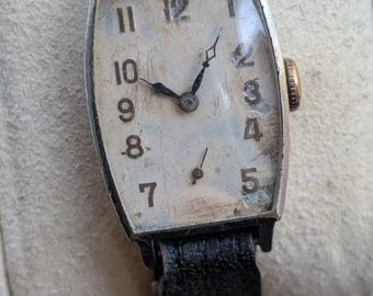 May include: A vintage wristwatch with a rectangular silver-tone case and a weathered off-white dial featuring black Arabic numerals. The watch has thin black hands and a small sub-dial at the bottom, attached to a worn black leather strap.