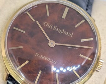 May include: A wristwatch with a gold-tone case and a dark brown, wood-grain patterned dial. The face features gold-tone hour markers, gold hands, and the text Old England and 17 JEWELS printed in gold. It includes a black leather watch strap.