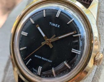 Könnte beinhalten: Eine goldfarbene Timex Armbanduhr mit rundem schwarzem Zifferblatt, weißen Stundenmarkierungen und Zeigern. Die Uhr hat ein strukturiertes dunkelbraunes Lederarmband. Auf dem Zifferblatt stehen die Wörter TIMEX und WATER RESISTANT.