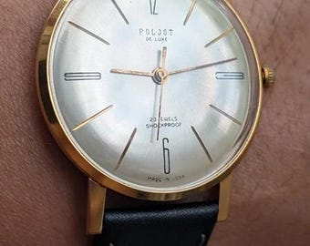 Könnte beinhalten: Eine goldfarbene analoge Armbanduhr mit rundem silbernem Zifferblatt, beschriftet mit POLJOT DE LUXE, 23 JEWELS SHOCKPROOF und MADE IN USSR. Die Uhr hat dünne goldfarbene Stundenmarkierungen, ein schwarzes Lederarmband mit Nähten und eine Krone.