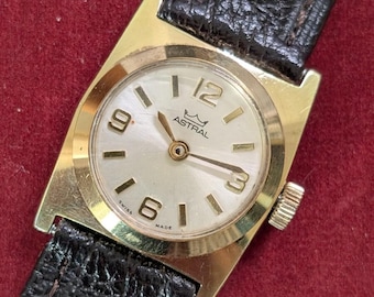 May include: A gold-tone wristwatch with a round, off-white dial featuring the text ASTRAL and a crown logo. The dial has gold-tone hour markers and numerals at 12, 3, 6, and 9 o'clock. It includes a textured black leather strap and Swiss Made text.