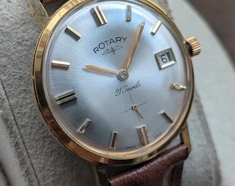 Könnte beinhalten: Eine goldfarbene Rotary Armbanduhr mit silbernem Sonnenschliff-Zifferblatt, goldenen Stundenmarkierungen und goldenen Zeigern. Die Uhr verfügt über ein Datumsfenster auf der Drei-Uhr-Position, den Schriftzug Rotary 21 Jewels und ein braunes Lederarmband.