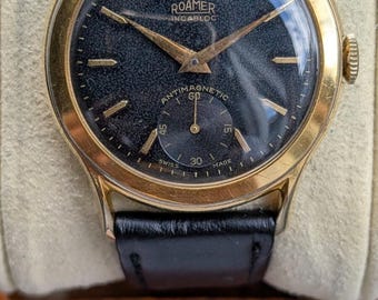 May include: A gold-tone wristwatch with a black textured dial, gold hands, and gold hour markers. The dial features the text ROAMER, INCABLOC, ANTIMAGNETIC, and SWISS MADE. It includes a sub-dial for seconds and a black leather strap.