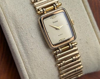 May include: A gold-tone Pulsar quartz wristwatch featuring a rectangular face with a champagne dial, gold hands, and a matching gold-tone metal link bracelet. The watch face displays the brand name Pulsar and the word Quartz in black text.