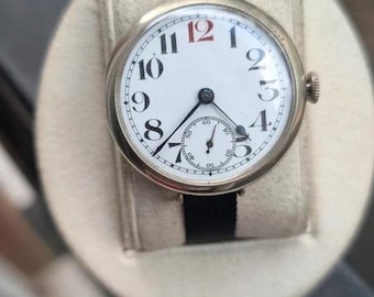 Könnte beinhalten: Eine runde Armbanduhr mit silberfarbenem Metallgehäuse und schwarzem Stoffarmband. Das weiße Zifferblatt zeigt schwarze arabische Ziffern, eine rote 12, schwarze Zeiger und ein kleines Hilfszifferblatt auf der Sechs-Uhr-Position für Sekunden.