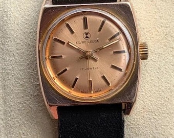 Könnte beinhalten: Eine Vintage-Armbanduhr von Favre-Leuba mit quadratischem goldfarbenem Gehäuse und rundem champagnerfarbenem Zifferblatt. Die Uhr verfügt über dünne goldfarbene Stundenmarkierungen, ein schwarzes Wildlederarmband und den Schriftzug Favre-Leuba 17 Jewels Swiss Made.