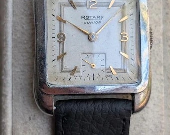 Könnte beinhalten: Eine rechteckige, silberfarbene Rotary Junior Armbanduhr mit weißem Zifferblatt, goldfarbenen Zeigern und goldfarbenen Ziffern auf den 12-, 3- und 9-Uhr-Positionen. Die Uhr verfügt über ein kleines Hilfszifferblatt für Sekunden und ein strukturiertes schwarzes Lederarmband.