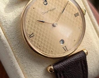 Könnte beinhalten: Eine goldfarbene Armbanduhr mit rundem Gehäuse, einem strukturierten goldenen Zifferblatt mit dem Courrèges-Logo und Schriftzug, römischen Ziffern bei drei, sechs und neun Uhr, einem Datumsfenster und einem dunkelbraunen Lederarmband mit Narbung.