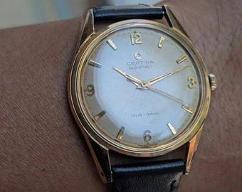 Könnte beinhalten: Eine goldfarbene Certina Automatic Blue Ribbon Armbanduhr mit silbernem Zifferblatt, goldfarbenen Zeigern und goldfarbenen Stundenmarkierungen, einschließlich der Ziffern 3, 6, 9 und 12. Die Uhr hat ein schwarzes Lederarmband mit sichtbaren Nähten.