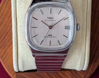 May include: A silver-toned Timex Automatic wristwatch with a rectangular case and a matching metal link bracelet. The watch features a light-coloured dial with thin hour markers, a date window at the three o'clock position, and the text Water Resistant.