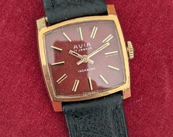 May include: A vintage Avia wristwatch featuring a square gold-tone case and a textured dark red dial. The watch has gold-tone hour markers and hands, with the text AVIA, 17 JEWELS, and INCABLOC printed on the face. It includes a black leather strap.