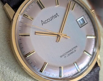 May include: A gold-tone wristwatch with a round, light-coloured dial featuring gold-tone hour markers and hands. The dial displays the text Accurist, SHOCKMASTER, and 17 JEWELS, with a date window at the three o'clock position and a dark brown strap.