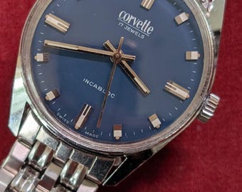 May include: A silver-tone analogue wristwatch with a round blue dial, featuring silver-tone hour markers and hands. The dial displays the text 'corvette', '17 JEWELS', 'INCABLOC', and 'SWISS MADE'. It includes a multi-link silver-tone metal bracelet.