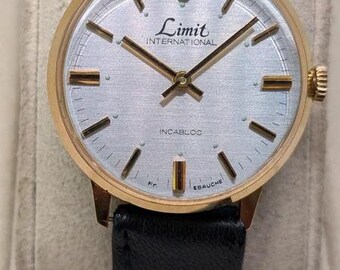 Könnte beinhalten: Eine goldfarbene Armbanduhr mit rundem silbernem Zifferblatt, goldenen Stundenmarkierungen und Zeigern. Auf dem Zifferblatt steht Limit INTERNATIONAL, INCABLOC und Fr. EBAUCHE. Sie hat ein schwarzes Lederarmband und eine seitliche Krone.