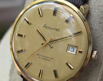 Könnte beinhalten: Eine Accurist Shockmaster Armbanduhr mit einem goldfarbenen runden Gehäuse, einem champagnerfarbenen Zifferblatt, goldfarbenen Stundenmarkierungen und Zeigern. Das Zifferblatt zeigt den Text Accurist, Shockmaster, 21 Jewels und ein Datumsfenster mit der 3.