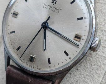 Könnte beinhalten: Eine Vintage-Armbanduhr von Junghans 17 Jewels mit silberfarbenem, rundem Zifferblatt, schwarzen Stunden- und Minutenzeigern sowie einem dünnen Sekundenzeiger. Die Uhr hat ein braunes Lederarmband und ein silberfarbenes Metallgehäuse mit Made in Germany.
