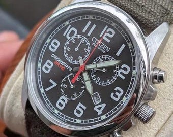 May include: A Citizen Eco-Drive chronograph watch with a black dial, large white Arabic numerals, and a red second hand. The stainless steel case features three sub-dials, a date window at the four o'clock position, and a textured olive green strap.
