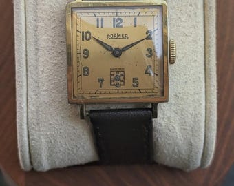 May include: A vintage square-cased Roamer wristwatch with a gold-toned dial, black Arabic numerals, and dark brown leather strap. The watch face features black hands, a small sub-dial at the six o'clock position, and the text ROAMER and SWISS MADE.