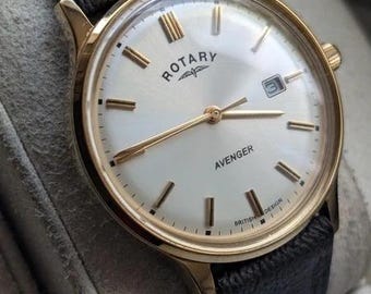 Könnte beinhalten: Eine goldfarbene Rotary Avenger Armbanduhr mit weißem Zifferblatt, goldenen Stundenmarkierungen, goldenen Zeigern und einem Datumsfenster auf der Drei-Uhr-Position. Die Uhr ist mit einem dunkelbraunen Lederarmband mit sichtbaren Nähten ausgestattet.