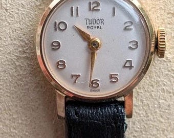 Könnte beinhalten: Eine Vintage Tudor Royal Armbanduhr mit einem runden goldfarbenen Gehäuse, einem weißen Zifferblatt mit schwarzen arabischen Ziffern und goldfarbenen Zeigern. Die Uhr ist mit einem strukturierten schwarzen Lederarmband und einer kleinen Krone ausgestattet.