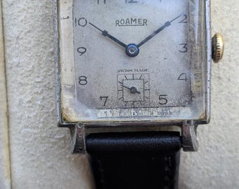 May include: A vintage Roamer wristwatch with a rectangular silver-toned case and a black leather strap. The off-white dial features black Arabic numerals, thin dark hands, and a small sub-dial at the six o'clock position with the text SWISS MADE.