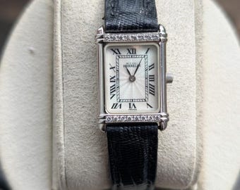 Könnte beinhalten: Eine rechteckige, silberfarbene Armbanduhr mit weißem Zifferblatt, schwarzen römischen Ziffern und dem Schriftzug Michel Herbelin France. Das Gehäuse ist oben und unten mit kleinen klaren Steinen besetzt und hat ein schwarzes, strukturiertes Lederarmband.