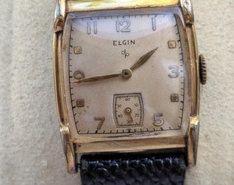 May include: A vintage Elgin wristwatch with a rectangular gold-tone case and a textured black leather strap. The off-white dial features gold-tone hands, Arabic numerals at 12, 7, and 5, square hour markers, and a small sub-dial for seconds.