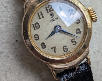 May include: A vintage Tudor wristwatch with a round gold-tone case, a cream-coloured dial featuring black Arabic numerals, and a black textured leather strap. The dial displays the Tudor logo and the word SWISS at the bottom with dark hands.