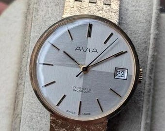 Könnte beinhalten: Eine Avia Armbanduhr mit einem goldfarbenen, strukturierten Metallgliederarmband und einem runden Gehäuse. Das Zifferblatt zeigt ein zweifarbiges silbernes Quadrantenmuster, dünne goldene Indizes, goldene Zeiger, ein Datumsfenster und Text.